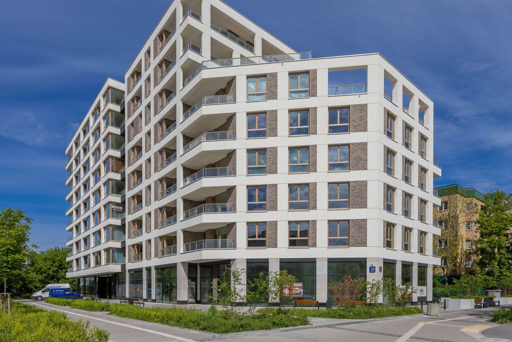 A modern residential building with a white facade, geometric balconies, and surrounding greenery.
