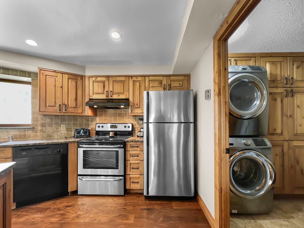 A kitchen with wooden cabinets, a stainless steel refrigerator, an oven, a dishwasher, and a small laundry area with a stacked washer and dryer.