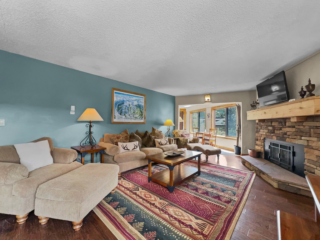 A cozy living room with a couch, armchair, and coffee table on a patterned rug. A stone fireplace sits to the right, and there is a large window and a door leading outside. A painting hangs on the wall.