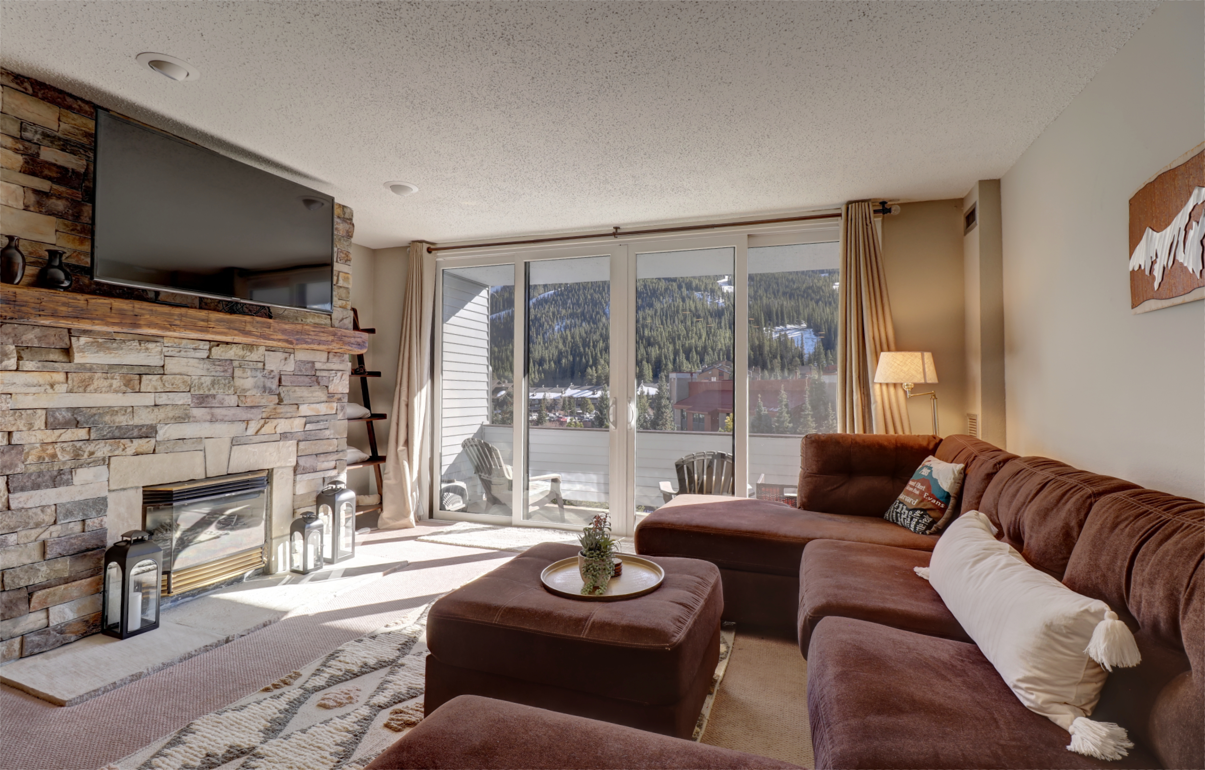 A cozy living room with a stone fireplace, a large wall-mounted TV, a brown sectional sofa, and large sliding glass doors opening to a balcony with a scenic mountain view.