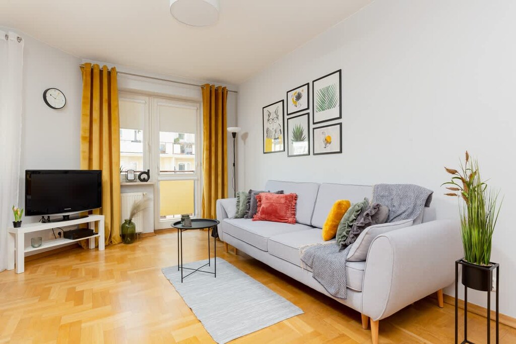 A bright and modern living room featuring a gray sofa, a small coffee table, and decorative wall art. The space is complemented by a green plant and natural light.