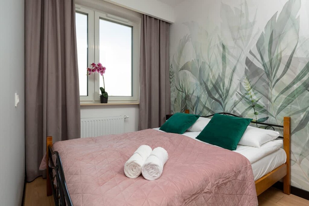 A bedroom with a large bed covered in pink and green accents, complemented by botanical wallpaper for a fresh and calming atmosphere.