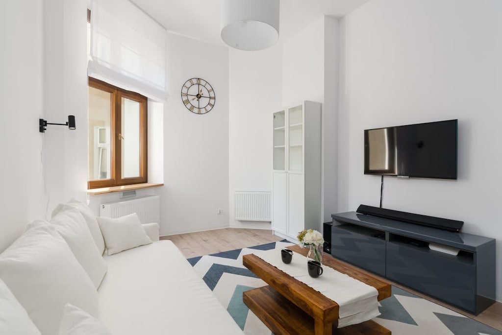 A bright, modern living room with a white sofa, wooden coffee table, and a TV mounted on the wall.