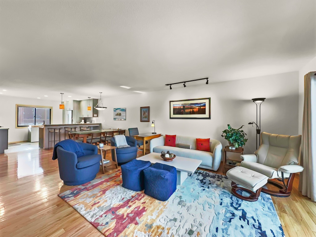 A modern living room with a light-colored sofa, blue and red accent chairs, a patterned rug, a floor lamp, and a wall-mounted framed artwork. The open kitchen and dining area are visible in the background.