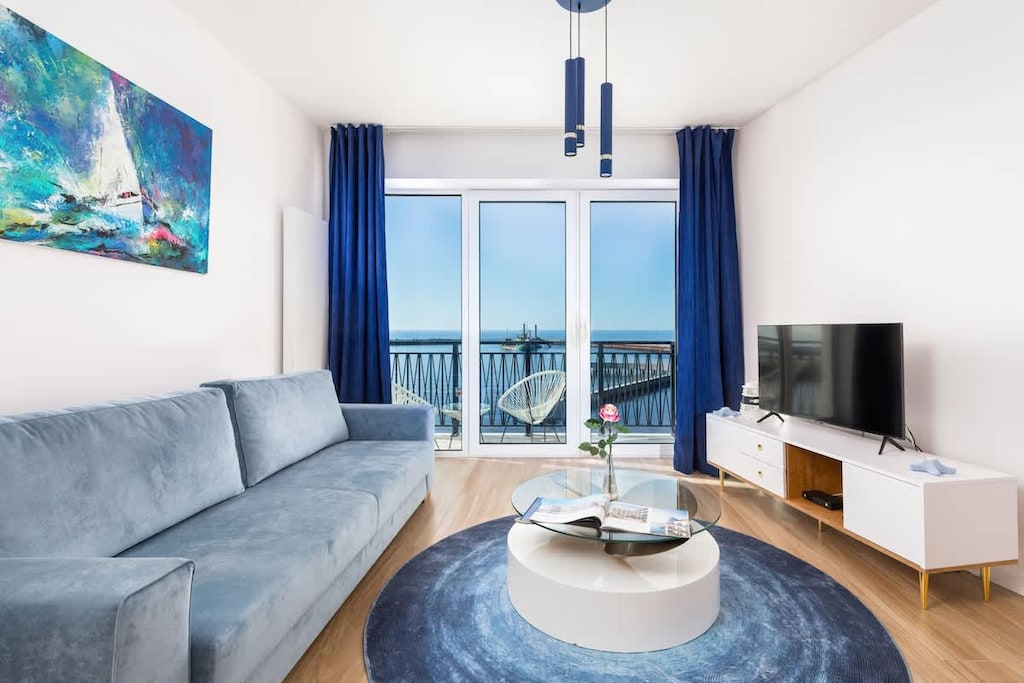 A modern living room with a blue and white color scheme, featuring a cozy gray sofa, a round coffee table, and a large window with stunning water views.

