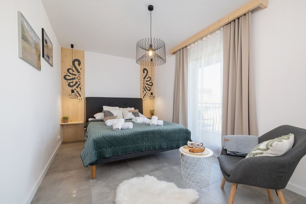 A bright living room featuring a gray sofa, a green rug, and a coffee table, with large windows letting in natural light.
