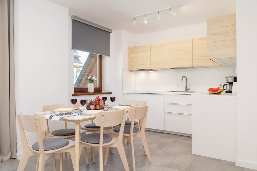 A modern kitchen with wooden cabinets, white countertops, and a dining area with a round table and chairs, blending functionality and style.

