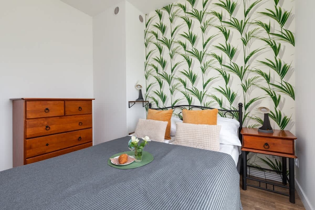 A cozy bedroom featuring a double bed with gray bedding, wooden furniture, and decorative wallpaper with a green leaf pattern.
