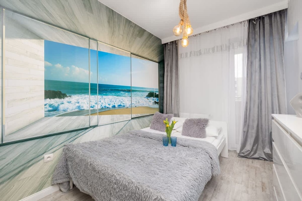 A cozy bedroom featuring a large mural of a beach, grey bedding, and light curtains.
