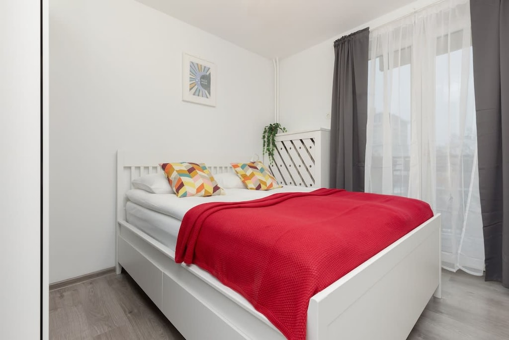 A bright bedroom featuring a double bed with a red blanket, a white desk, and large windows for natural light.