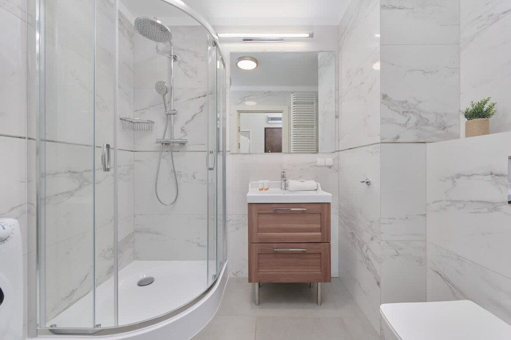 A modern bathroom with a glass-enclosed shower cabin, marble walls, and a wooden vanity with a white basin, offering a clean and luxurious feel.
