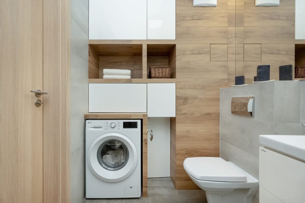 A compact bathroom with light wood and white tiles, featuring a washing machine, toilet, and minimalist storage solutions.
