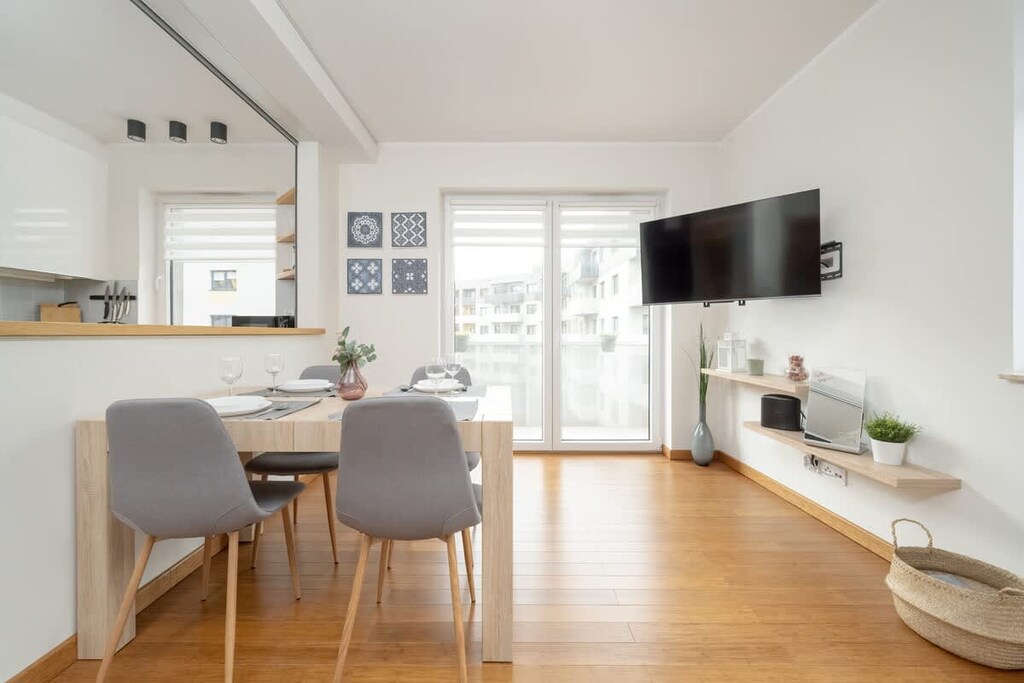 An open-plan living and dining area with a table set for two, grey chairs, and a TV mounted on the wall.
