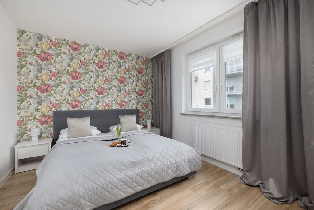 A cozy bedroom with a soft bed, floral-patterned wallpaper, elegant curtains, and a warm, inviting atmosphere.

