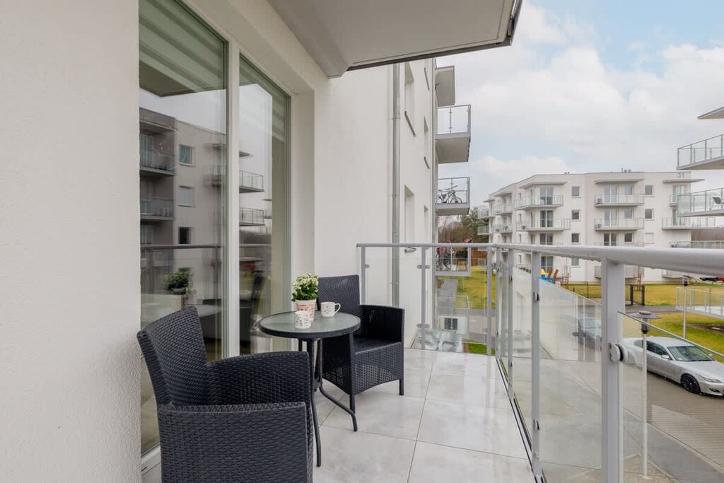 A spacious balcony with outdoor seating, overlooking a modern residential complex with glass railings for an open feel.

