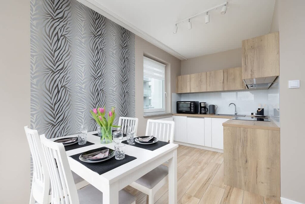 A bright dining area next to the kitchen, featuring a white table, black tableware, and large windows bringing in natural light.

