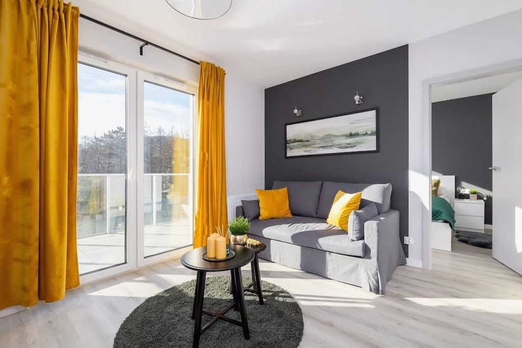A modern living room with a dark accent wall, a gray sofa, mustard yellow curtains, and a round coffee table, creating a stylish and cozy atmosphere.

