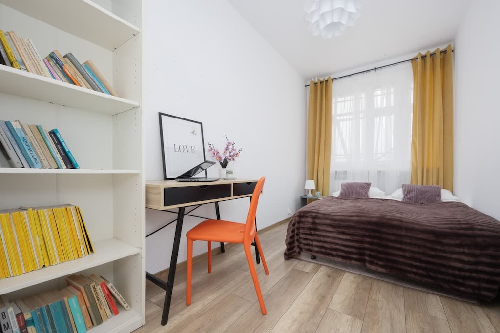 A bedroom featuring a brown bedspread, white wardrobe, and a shelf with colorful books and decor.

