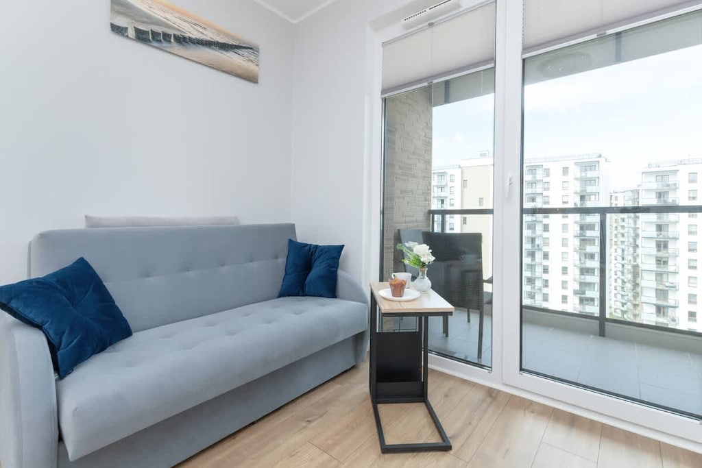 A bright living room with a blue sofa and large windows opening to a balcony. Natural light fills the room, creating a peaceful atmosphere.