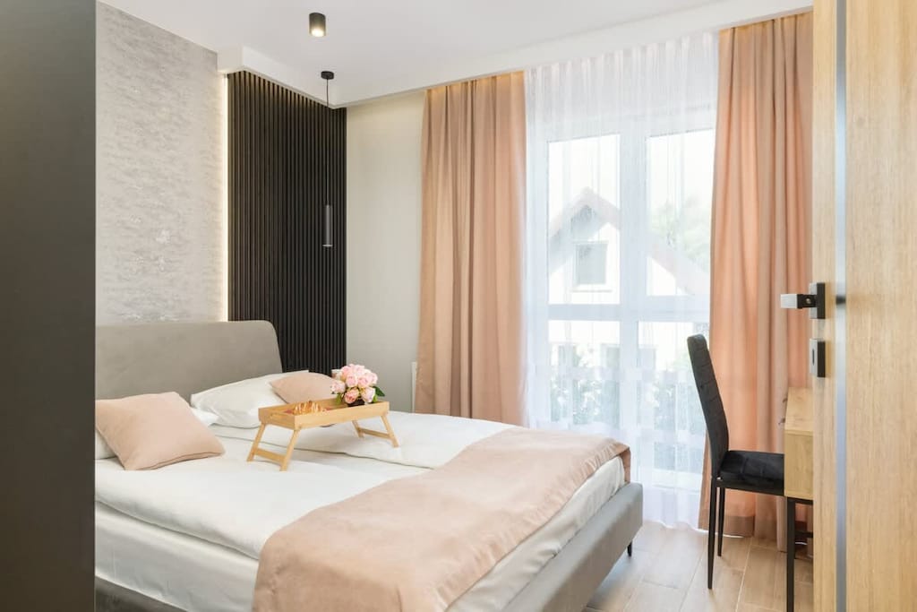 A cozy bedroom with a large bed and sliding wardrobe doors. Natural light pours in through sheer curtains.