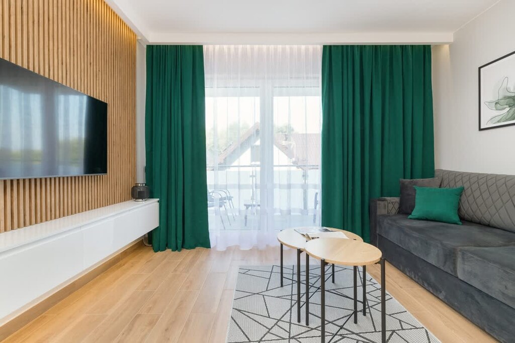 A contemporary living room with green curtains and wooden accents. The space feels airy and vibrant.