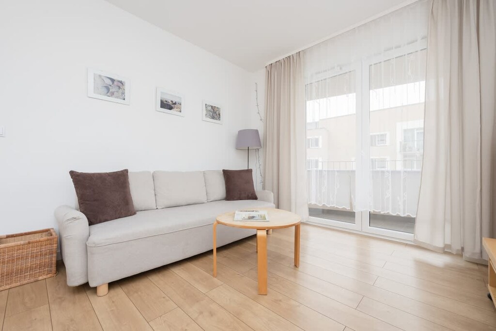 A bright living room with a beige sofa, a wooden coffee table, and sheer curtains allowing natural light to flood the space.