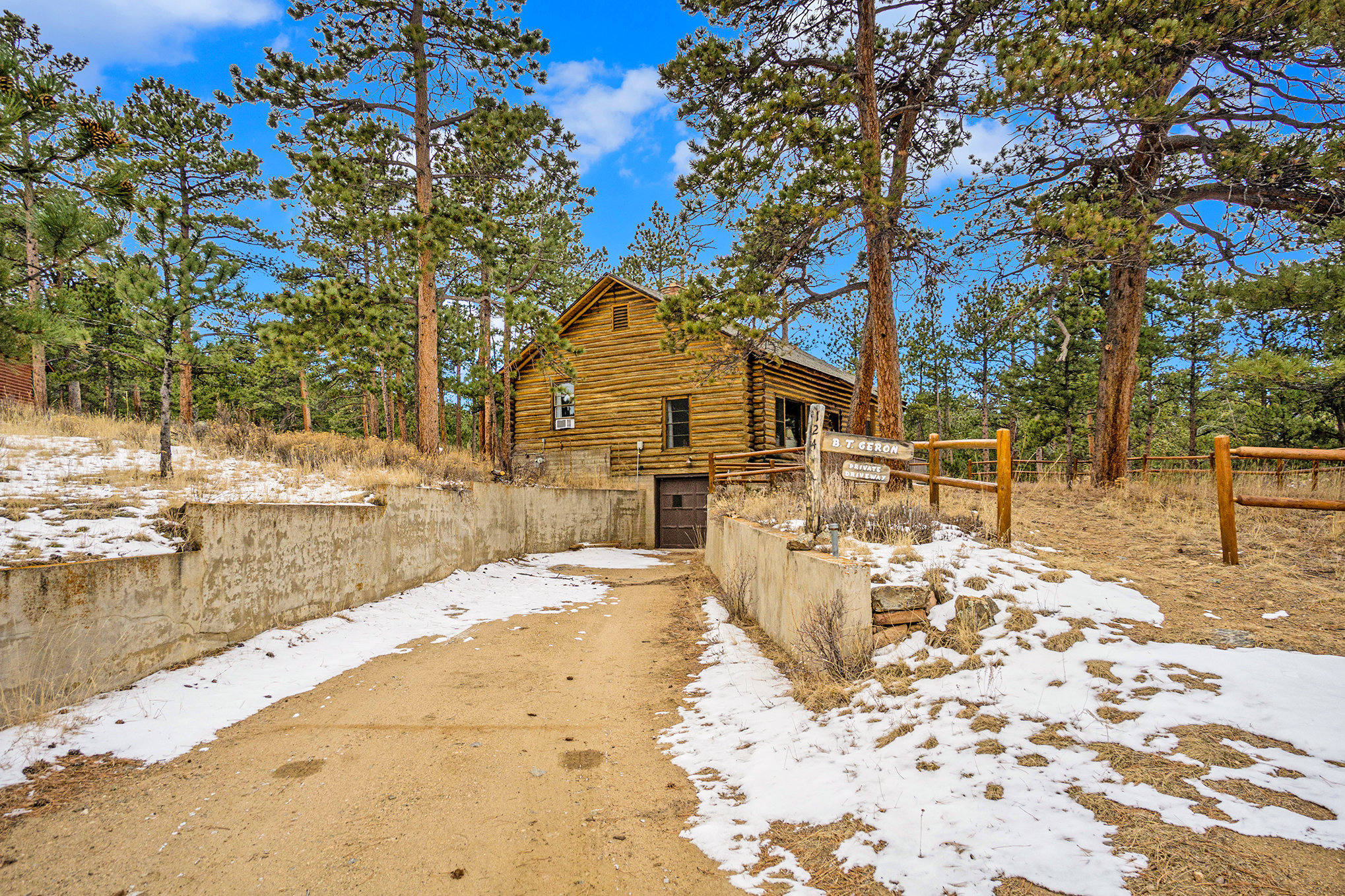 A rustic wooden cabin surrounded by pine trees, with a snowy ground and a paved path leading to its entrance.