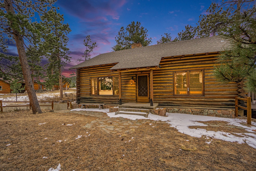 Log cabin surrounded by trees with light snow on the ground, set against a colorful twilight sky.