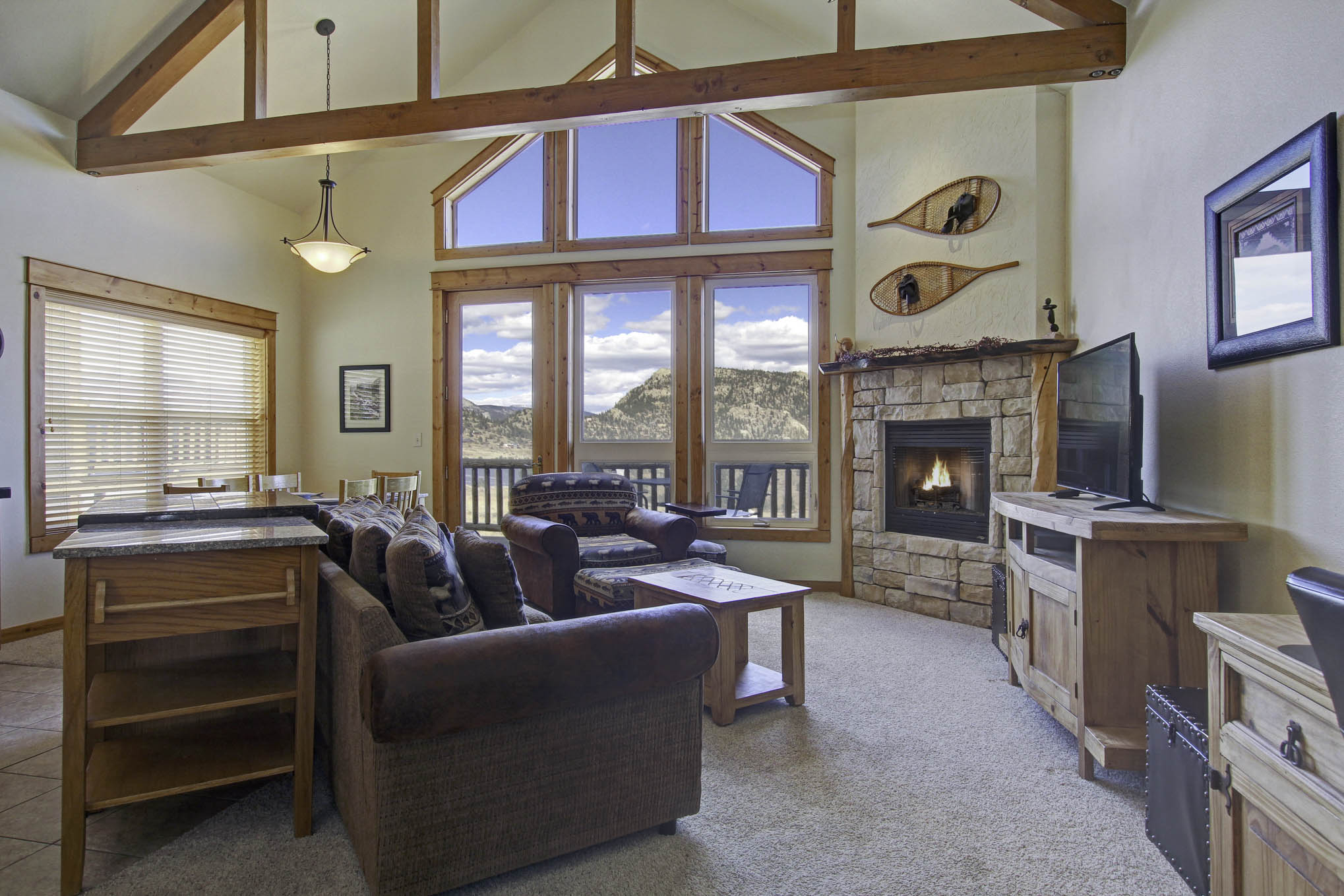 Condo's main living area boasts floor to ceiling windows with stunning mountain views, a stone fireplace, flat screen TV, and ample seating from either the double sofa or loveseat. Opens up to the dining/kitchen area.