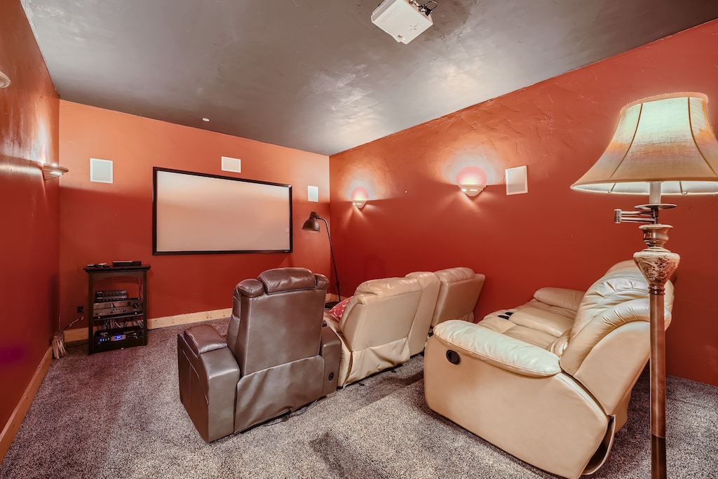 A home theater room with red walls, a large screen, a projector, and several recliner chairs arranged in rows. A floor lamp is visible to the right.