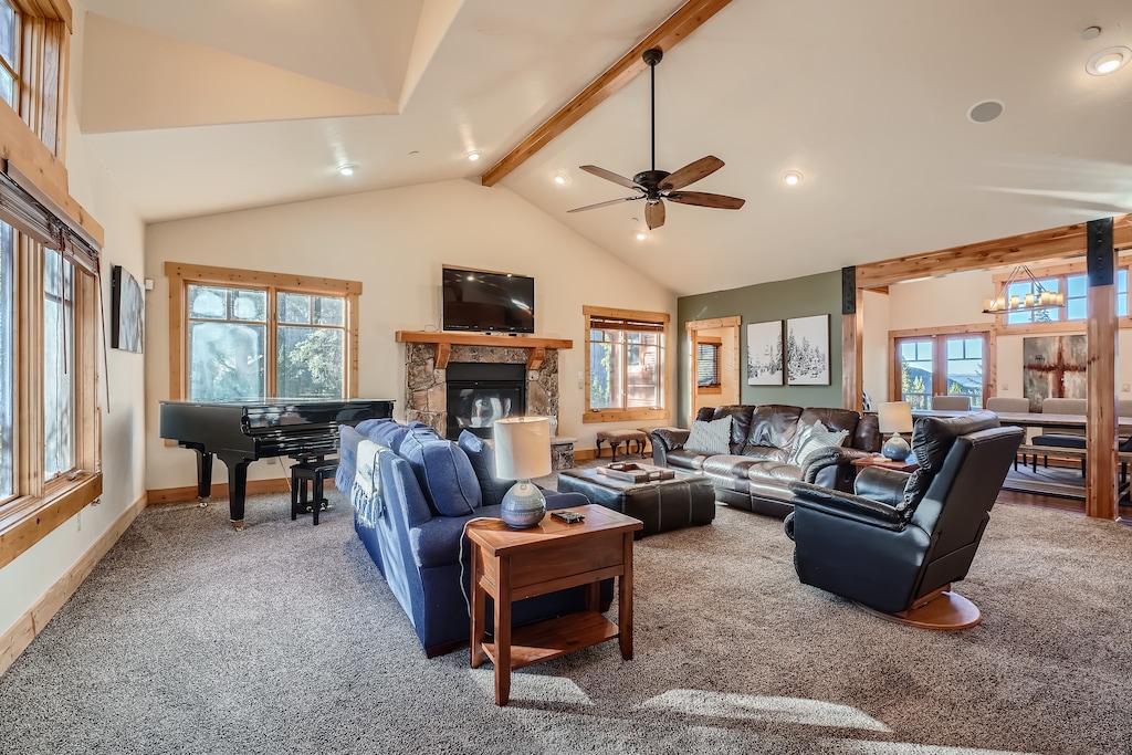 A spacious living room with a vaulted ceiling, furnished with sofas, recliner, a coffee table, and a grand piano. A ceiling fan and a fireplace with a TV above complete the cozy setup.