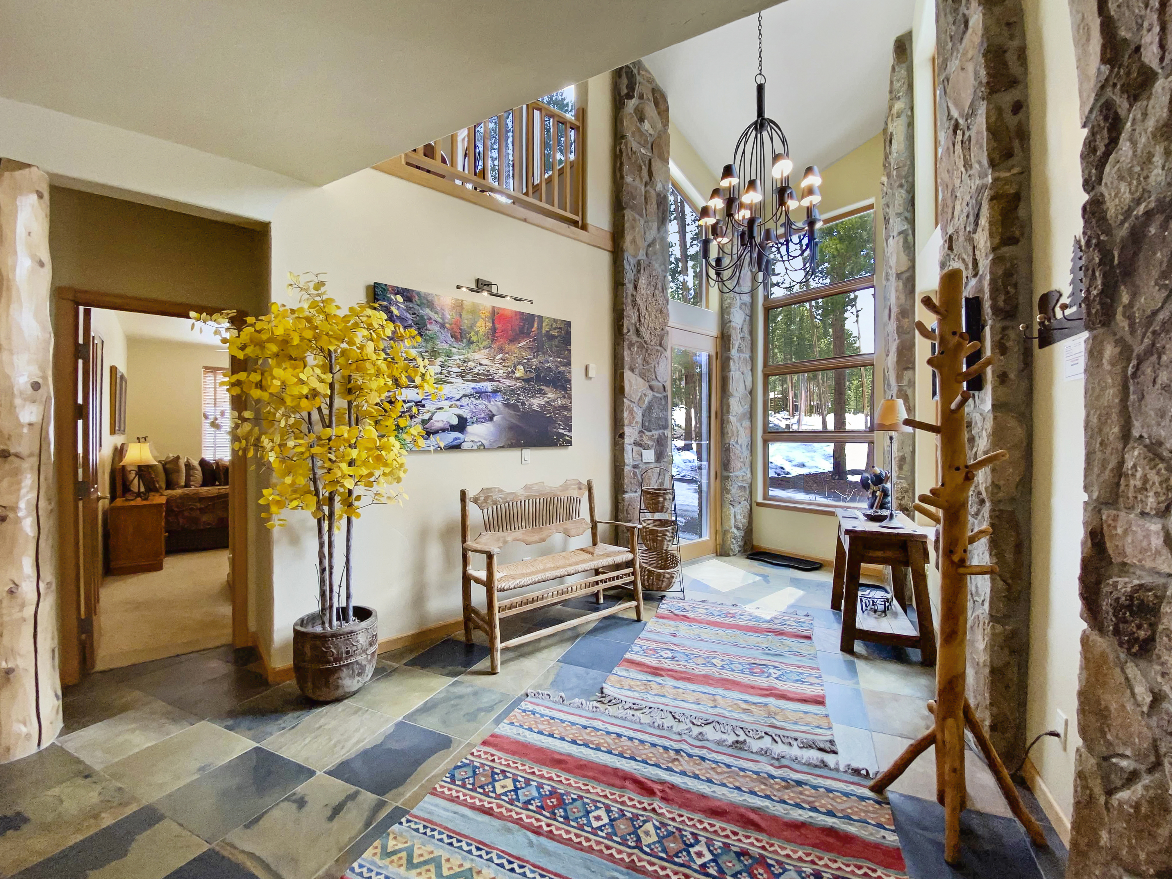 A brightly lit entryway with a multicolored rug, wooden bench, potted plant, and wall art. Stone pillars and chandelier enhance the rustic decor. Stairs and a living room are visible in the background.