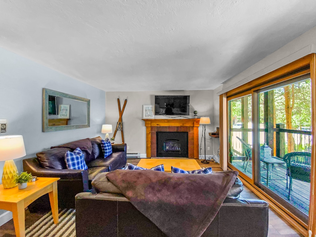A cozy living room featuring a fireplace, leather sofas with plaid cushions, a wall-mounted TV, large mirror, and sliding glass doors leading to an outdoor patio with wicker chairs.