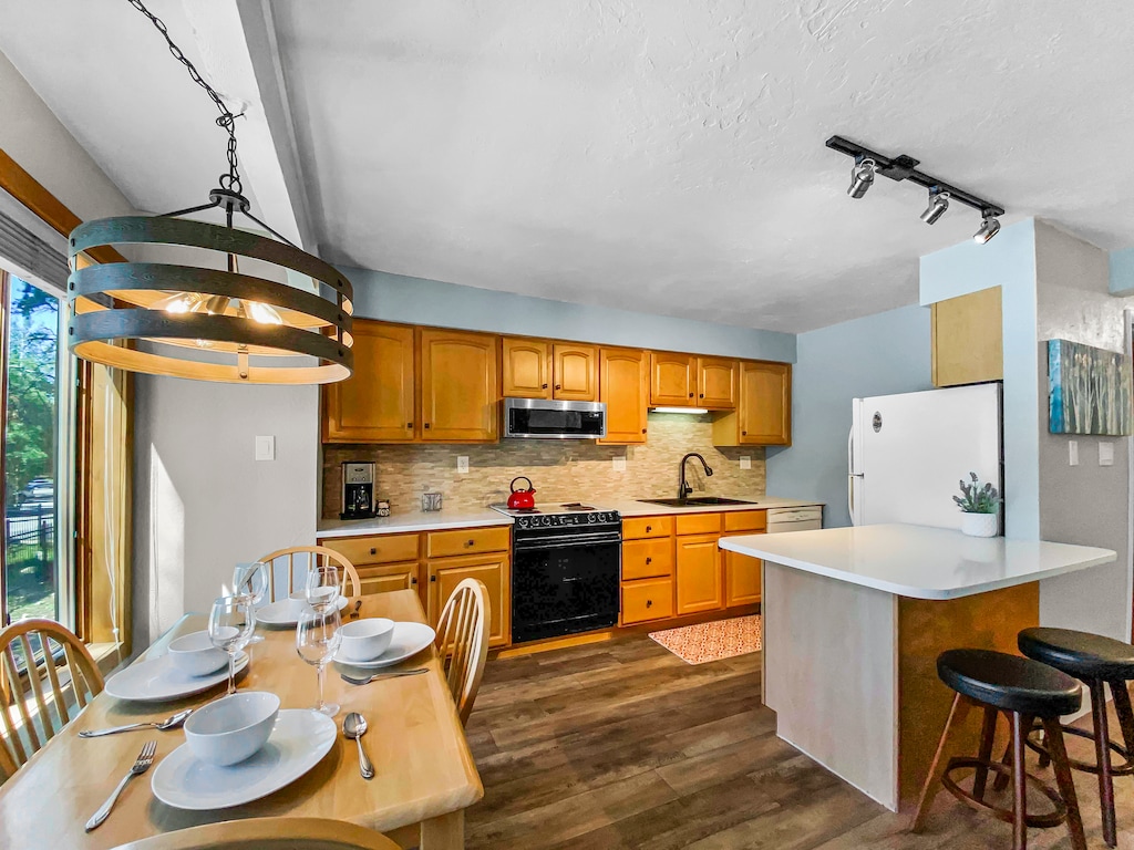 A well-lit kitchen with wooden cabinets, a black oven, a white refrigerator, a countertop bar with two stools, and a dining table set with dishes and cutlery. A window is visible on the left.