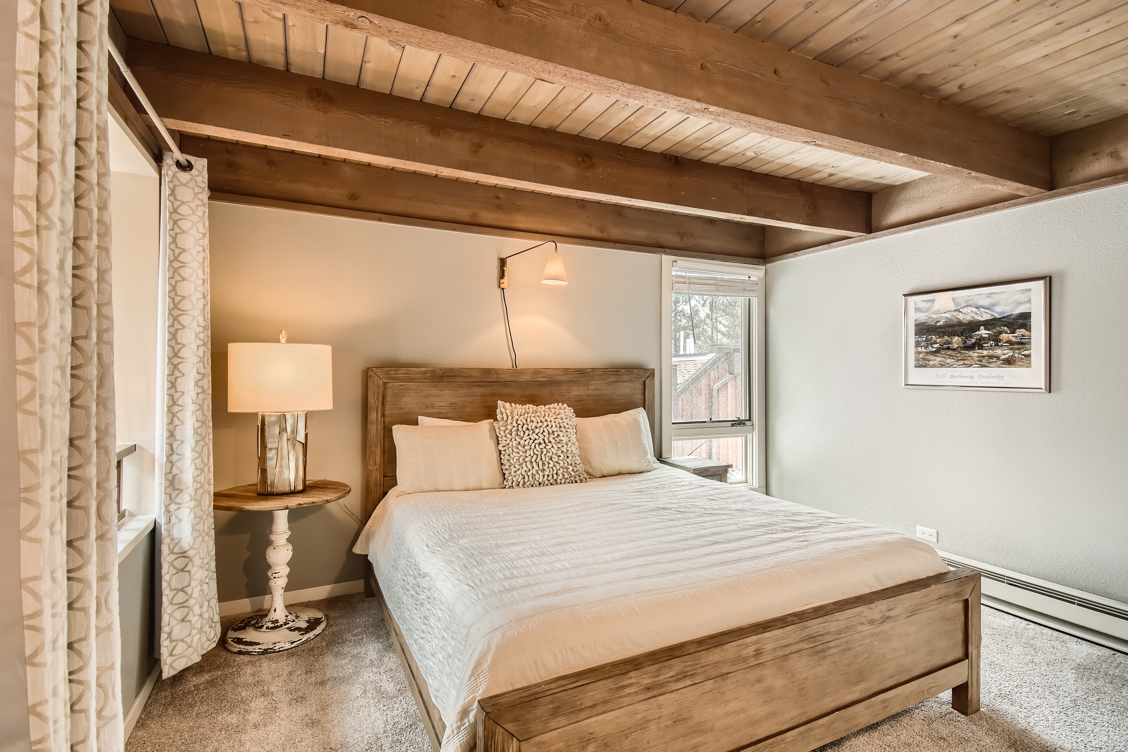 A cozy bedroom with a wooden bed frame, white bedding, a lamp on a side table, a wall-mounted reading light, and a window with curtains. Exposed wooden ceiling beams and a framed picture on the wall.