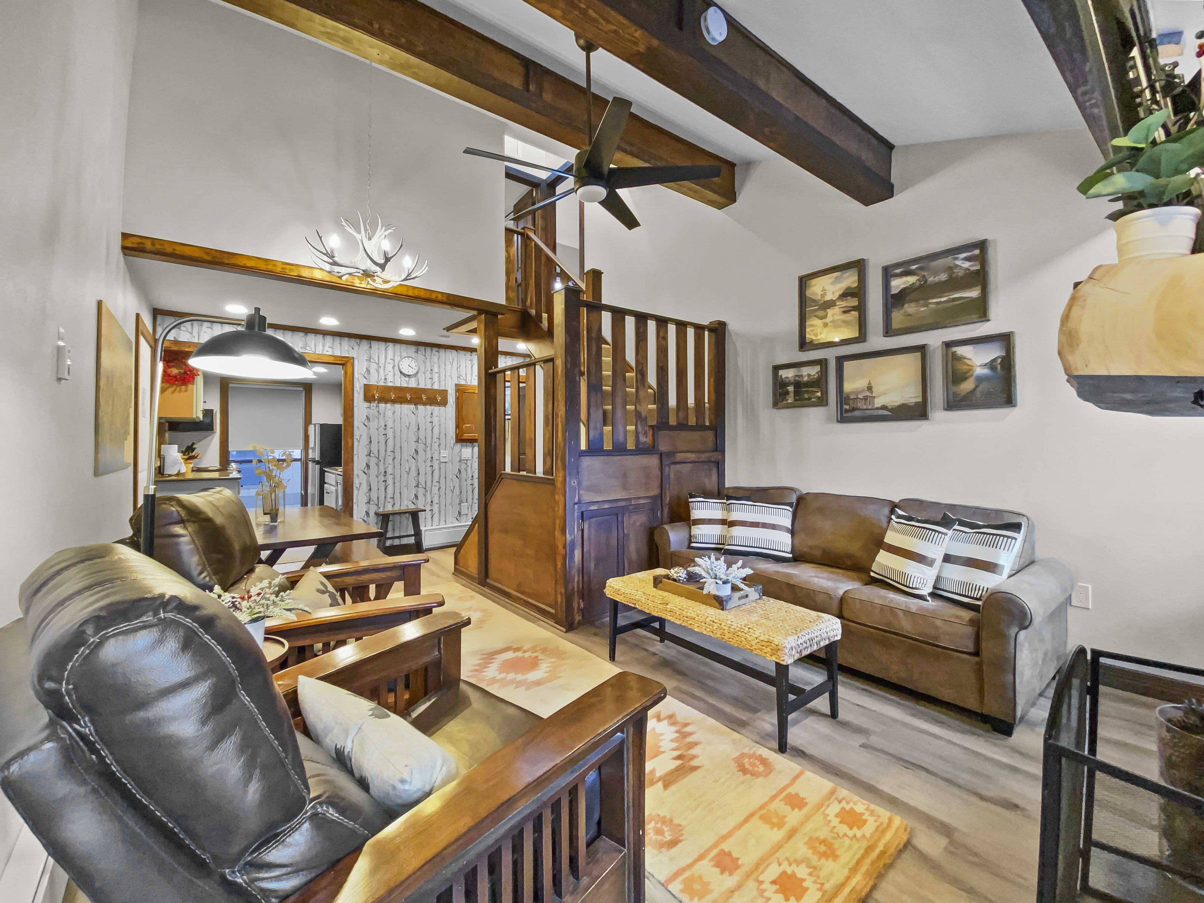 A cozy living room with wooden beams, leather sofas, armchairs, a ceiling fan, framed pictures on walls, and a staircase leading to a loft. Warm lighting highlights the rustic decor.