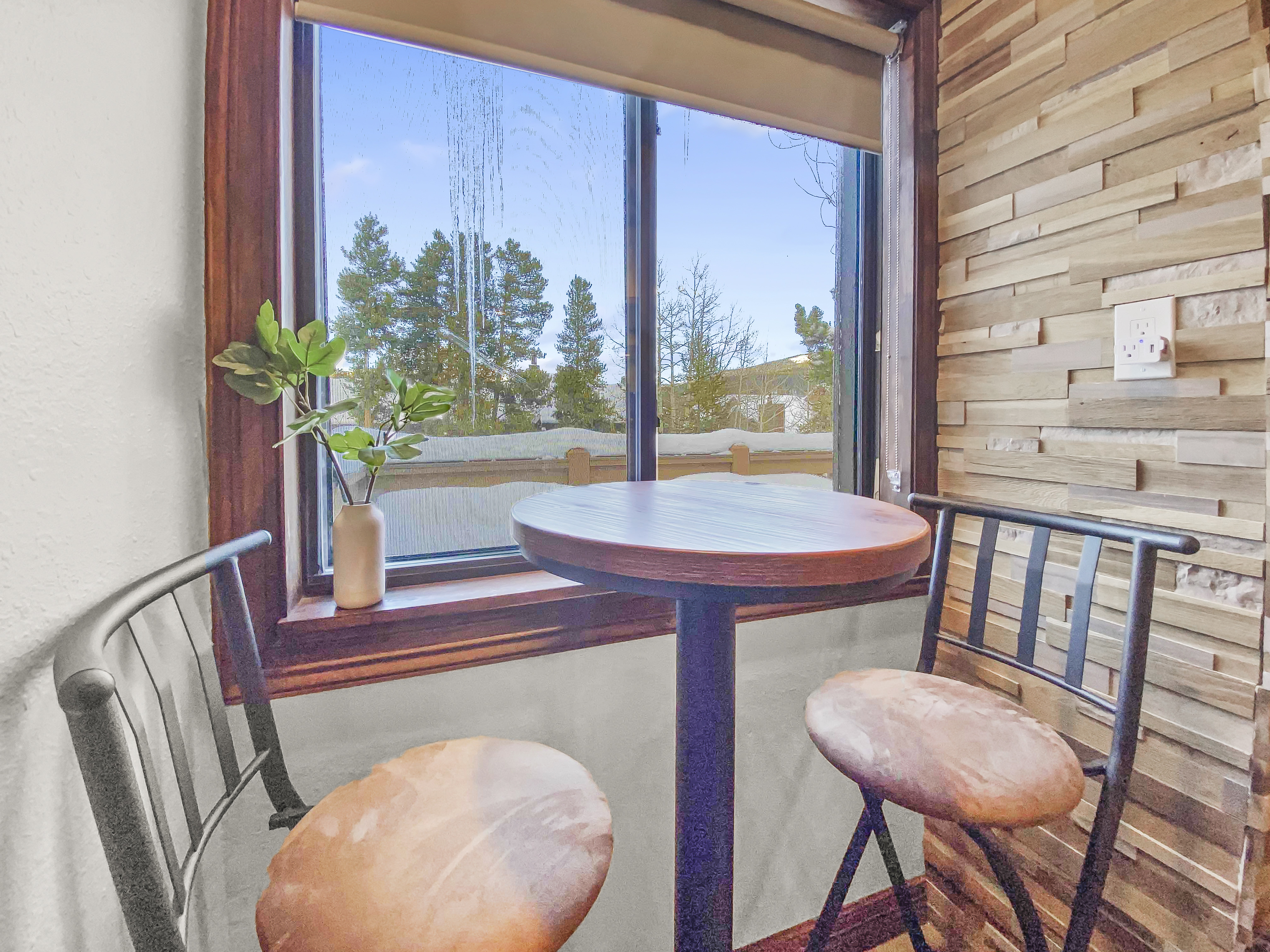 A small, round table with two chairs is placed next to a window overlooking trees and a building. The setting features a beige, textured wall and a tiny plant on the table.