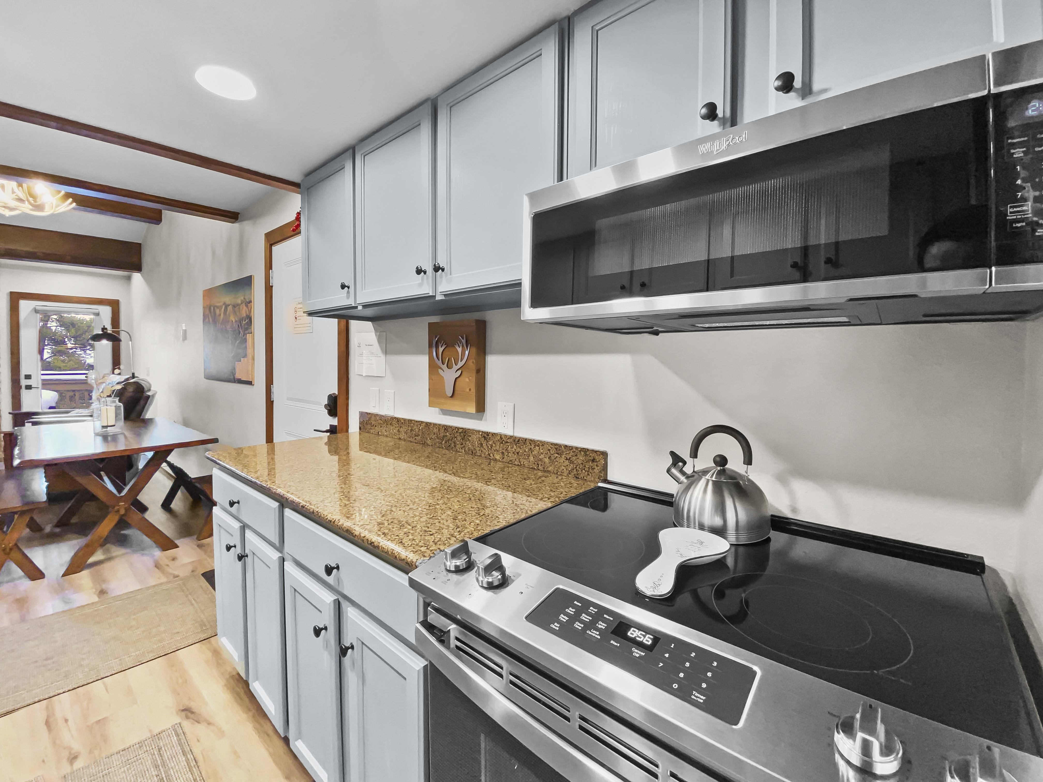 A kitchen with light blue cabinets, granite countertops, a stainless-steel stove with overhead microwave, and a kettle on the stovetop. A dining table is visible in the adjoining room.