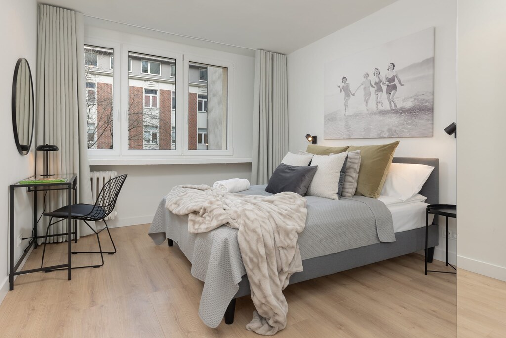 A bedroom setup with large windows, cozy bedding, and a blanket draped over a chair.
