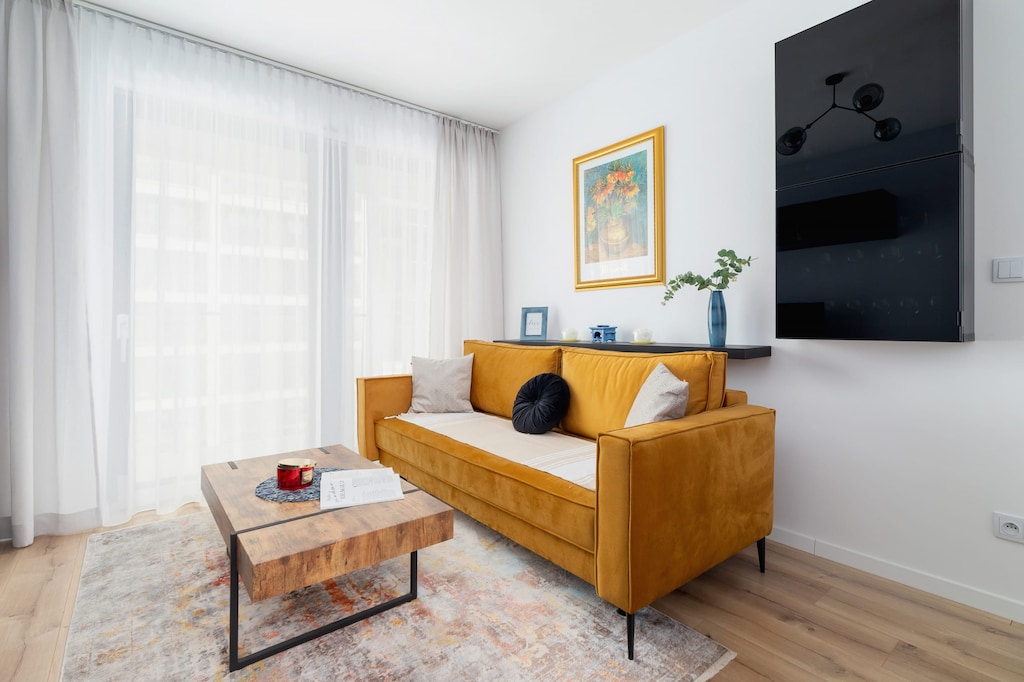A bright living room with a mustard yellow sofa, a wall-mounted TV, and large windows.
