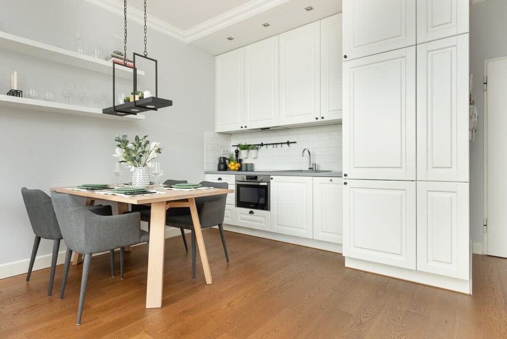 An open kitchen and dining area in white, featuring a wooden table, pendant lights, and ample storage.
