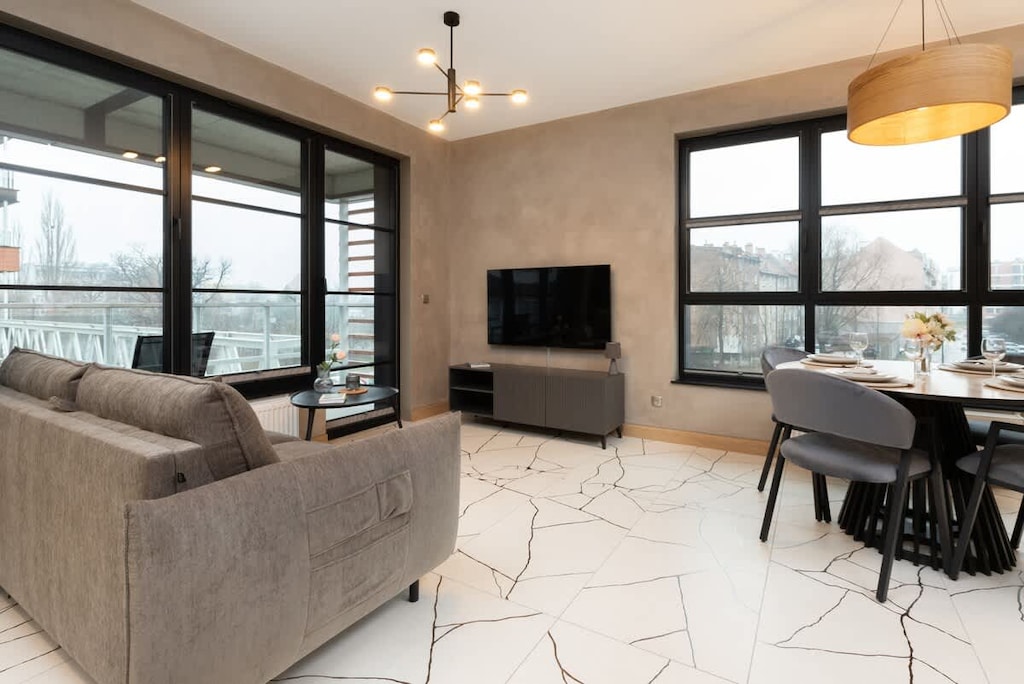 A modern living room with large windows, a comfortable gray sofa, a TV on a sleek cabinet, and a stylish dining area in the background.

