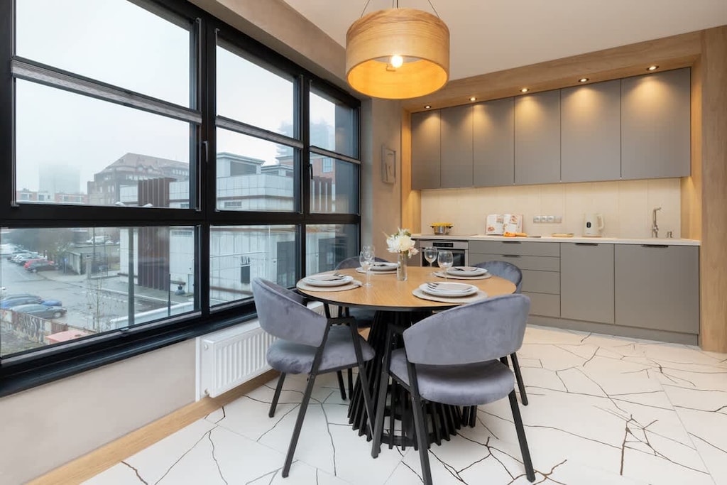 A sophisticated dining space with a round table, plush gray chairs, a pendant light above, and panoramic windows providing a city view.

