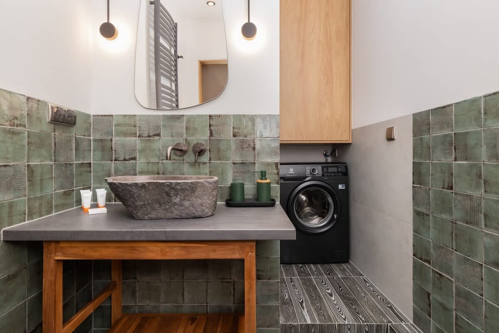 A contemporary kitchen corner with green tiled walls, wooden cabinets, a washing machine, and a stone-textured sink for a unique aesthetic.

