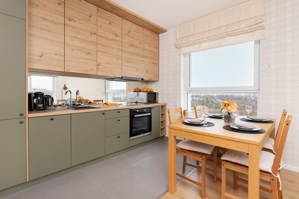 A stylish kitchen with wooden upper cabinets, green lower cabinets, and a small dining area by the window, creating a warm and functional space.

