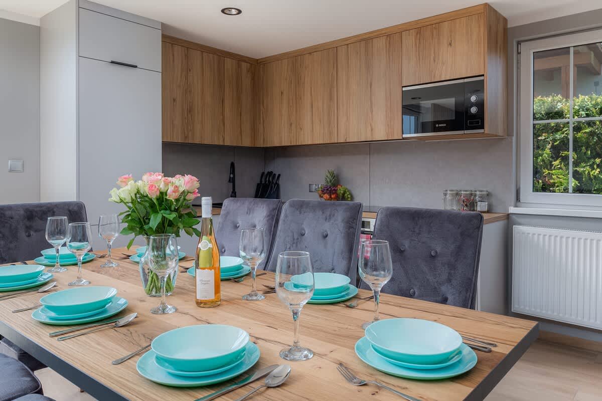 A spacious kitchen-dining area with a wooden dining table, set with turquoise plates and glasses, overlooking a fully equipped kitchen in the background.