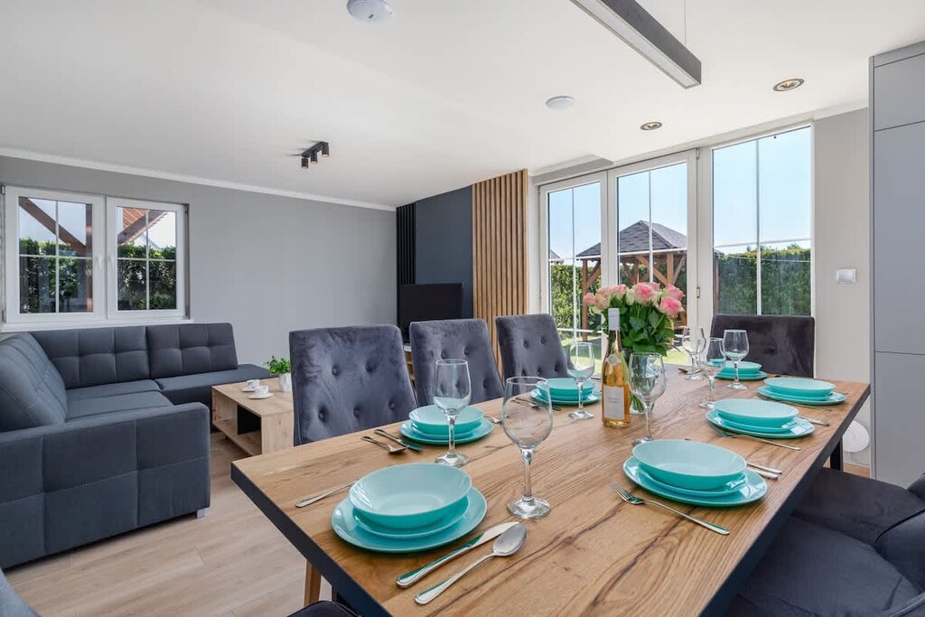 A spacious kitchen-dining area with a wooden dining table, set with turquoise plates and glasses, overlooking a fully equipped kitchen in the background.