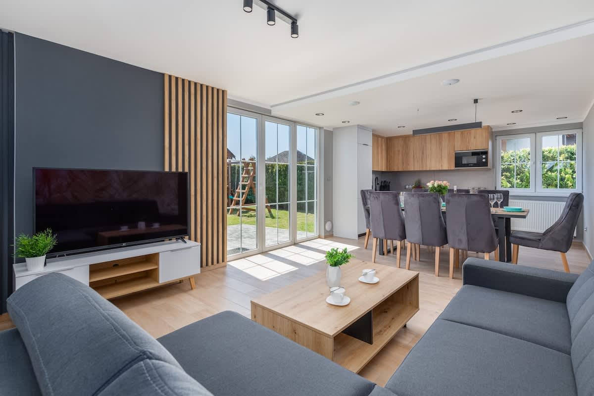 A bright, modern living room featuring a gray sofa, a light wooden coffee table, and large windows that let in plenty of natural light, with a garden view in the background.