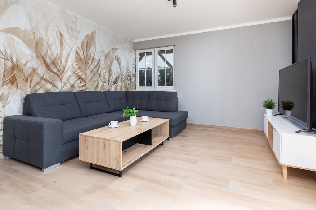A bright, modern living room featuring a gray sofa, a light wooden coffee table, and large windows that let in plenty of natural light, with a garden view in the background.