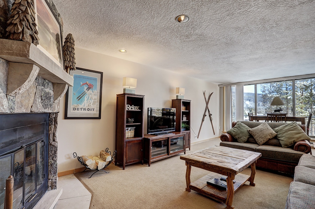 A cozy living room with a stone fireplace, wood furniture, a TV, skis on the wall, and a large window offering a scenic view. A framed "Detroit" poster is displayed above the fireplace.